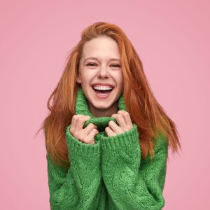 Modern ginger woman cuddling in cozy green sweater and laughing at camera on pink background